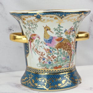 May include: A porcelain vase featuring a central illustration of a pheasant surrounded by colorful foliage. The vase has a flared rim, two gold-toned handles, and a deep blue base with intricate gold filigree patterns and a scalloped border.