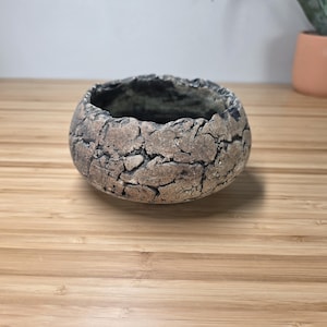 May include: A small, round, textured ceramic bowl with a cracked, stone-like appearance. The bowl is primarily shades of brown and gray, with a dark interior. The bowl sits on a light brown wooden surface. A terracotta pot with a plant is in the background.
