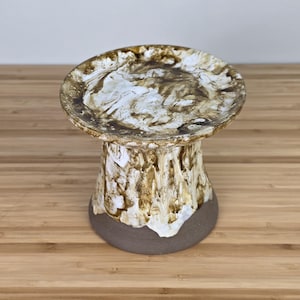 May include: A ceramic pedestal with a round top and a flared base. The top features a marbled pattern of brown and white, while the body has a textured, bark-like appearance with brown and white tones. The base is a solid, dark gray color.