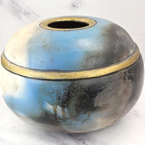 May include: A round, low-profile ceramic vessel featuring a smooth, matte finish with a gradient of blue, black, and off-white tones. The piece is accented by a horizontal gold-colored band around the center and a matching gold-colored rim.