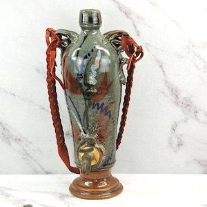 May include: A tall, glazed ceramic vessel with a narrow neck and a wide base. The body is mottled green with abstract blue and reddish-brown painted accents. A metal spigot is attached near the bottom, and two braided rust-colored leather straps are attached to the handles on either side of the neck.