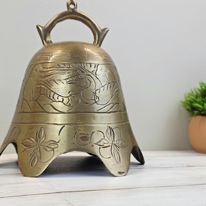 May include: A brass bell featuring an arched handle at the top and a scalloped bottom edge. The surface is decorated with engraved patterns, including a central motif of a bird in flight and floral designs around the base of the bell.
