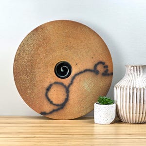 May include: A large, round, ceramic wall art piece with a textured, tan surface and a black spiral design. A small, white speckled planter with a succulent and a white and tan striped vase are next to it.