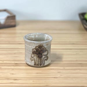 May include: A small, cylindrical ceramic cup with a beige glaze. The cup features a brown floral design on the front. The cup is sitting on a light brown wooden surface.