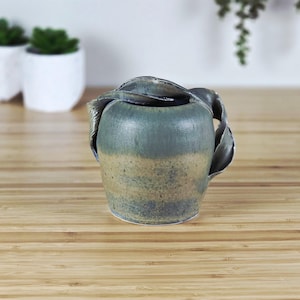 May include: A handmade ceramic vase with a unique design. The vase is a blend of blue and tan hues, with a textured surface. It features a decorative, leaf-like handle. The vase is approximately 5 inches tall.