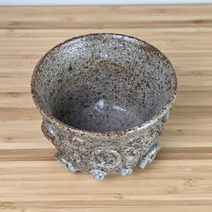 May include: A speckled ceramic bowl with a textured surface and a rounded shape. The bowl is primarily gray with brown speckles and decorative, raised circular designs around the base. The bowl sits on a light brown wooden surface.
