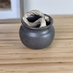 May include: A small, round, dark gray ceramic pot with a textured surface. The pot contains a light-colored, twisted, organic-looking object. The pot sits on a light brown wooden surface. Decorative pottery.