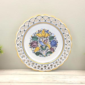 May include: A round ceramic plate with a scalloped, yellow-rimmed edge featuring a lattice-style cutout border. The center displays a hand-painted floral motif with a central blue bird surrounded by yellow, purple, and green stylized flowers.