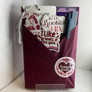 May include: A burgundy gift set for book lovers, featuring stickers with text like "In My Bookish Era" and "I like my books extra spicy." Includes a bookmark with a tassel and a pen. A sticker with "Romance Reader" is also visible.