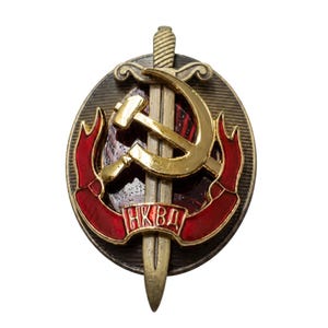 May include: An oval-shaped metal badge featuring a vertical sword overlaid with a gold-colored hammer and sickle emblem. A red enameled ribbon curves across the lower section, displaying the Cyrillic text NKVD in gold lettering against a dark background.