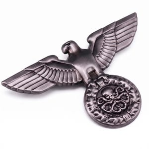 May include: A metallic gunmetal gray eagle emblem featuring outstretched wings. The eagle is perched atop a circular wreath containing a central skull motif with tentacle-like appendages extending from the base, rendered in a dark, polished finish.