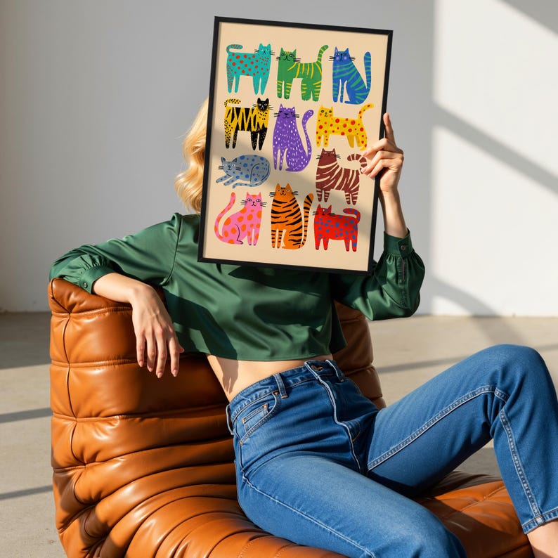 May include: A framed art print featuring a collection of colorful cat illustrations. The cats are depicted in various patterns and colors, including blue, green, yellow, and orange, against a beige background. The print is held by a person wearing a green top and blue jeans.