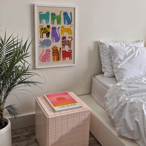 May include: A framed art print featuring a colorful pattern of stylized cats in various poses and colors. The artwork is displayed on a neutral wall above a pink tiled side table with a book on top. A potted plant and a bed with white linens are also visible.