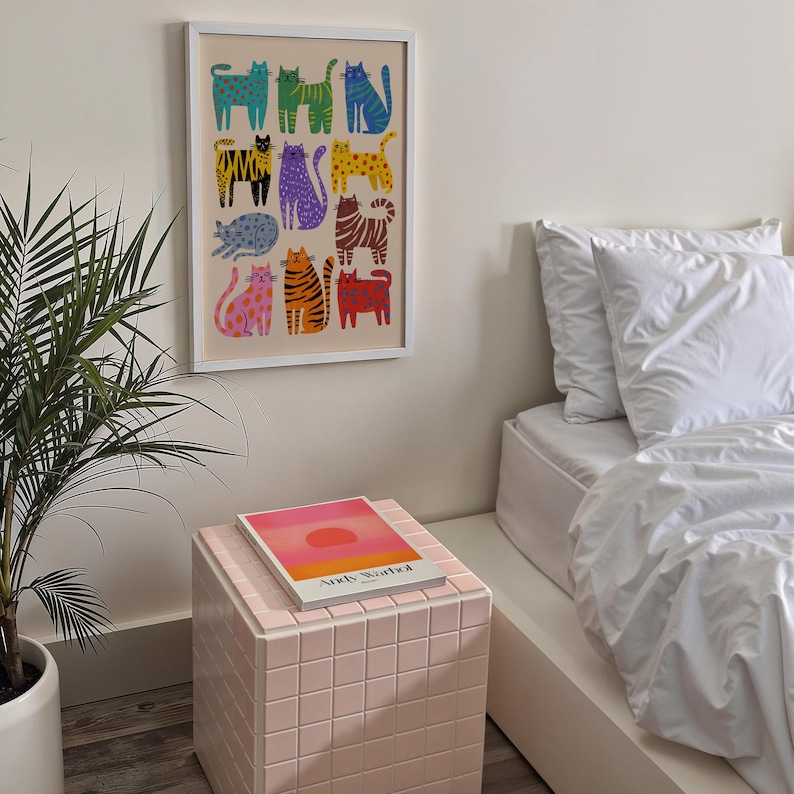 May include: A framed art print featuring a colorful pattern of stylized cats in various poses and colors. The artwork is displayed on a neutral wall above a pink tiled side table with a book on top. A potted plant and a bed with white linens are also visible.