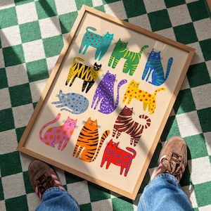May include: A framed art print featuring a collection of colorful, patterned cat illustrations. The cats are depicted in various poses and colors, including blue, yellow, orange, and purple, with patterns like polka dots and stripes. The artwork is set against a cream background.