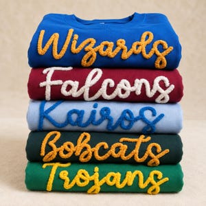 Könnte beinhalten: Ein Stapel aus fünf Rundhals-Sweatshirts in Blau, Kastanienbraun, Hellblau, Dunkelgrün und Grün. Jedes Sweatshirt hat eine strukturierte, erhabene Chenille-Schrift in Schreibschrift mit den Wörtern Wizards, Falcons, Kairos, Bobcats und Trojans.