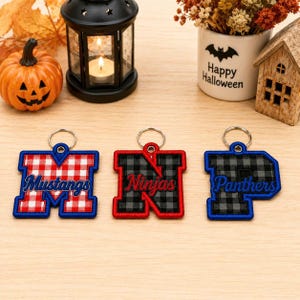 May include: Three embroidered letter keychains with metal rings. The first is a red and white buffalo plaid M with the word Mustangs. The second is a black and grey buffalo plaid N with the word Ninjas. The third is a black and grey buffalo plaid P with the word Panthers.