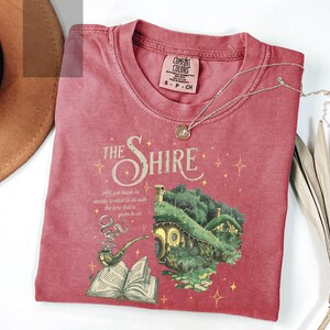 May include: A red t-shirt with a graphic of a house with a green roof and a book with a pipe. The text "The Shire" is above the house and the text "All we have to decide is what to do with the time that is given to us" is below the house.