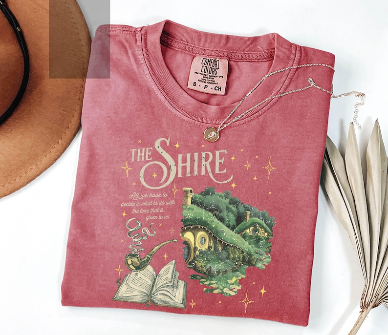 May include: A red t-shirt with a graphic of a house with a green roof and a book with a pipe. The text "The Shire" is above the house and the text "All we have to decide is what to do with the time that is given to us" is below the house.