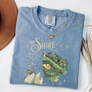 May include: A light blue t-shirt with a graphic of a hobbit house, a pipe, and an open book. The text "The Shire" is printed in yellow above the graphic. The quote "All we have to decide is what to do with the time that is given to us" is printed below the graphic.