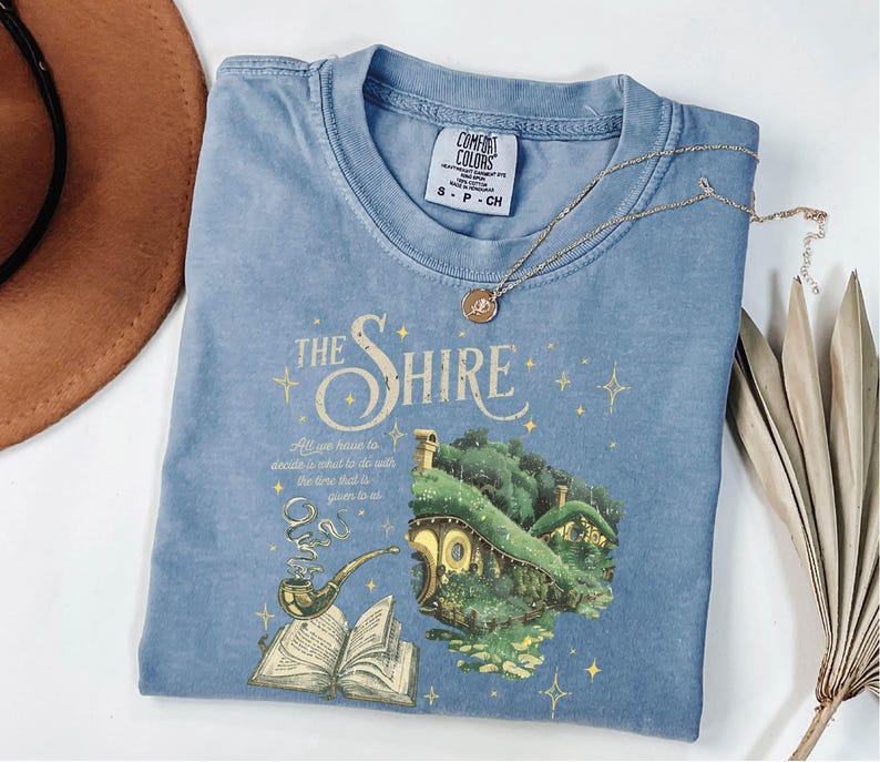 May include: A light blue t-shirt with a graphic of a hobbit house, a pipe, and an open book. The text "The Shire" is printed in yellow above the graphic. The quote "All we have to decide is what to do with the time that is given to us" is printed below the graphic.