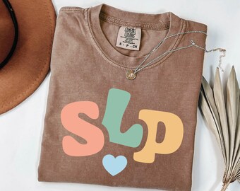 May include: A brown Comfort Colors t-shirt with the letters "SLP" in coral, teal, and yellow, and a light blue heart. A silver necklace with a small pendant is on the shirt.