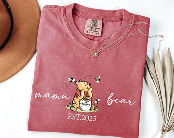 May include: A red t-shirt with a white graphic of Winnie the Pooh holding a honey pot. The graphic also includes bees and the text "mama bear EST.2025".