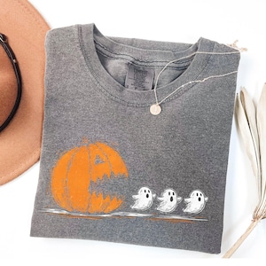 May include: A gray Comfort Colors t-shirt featuring an orange pumpkin with a carved-out face and three white ghost illustrations. A silver necklace with a round pendant is on the shirt. A brown hat is in the upper left corner.