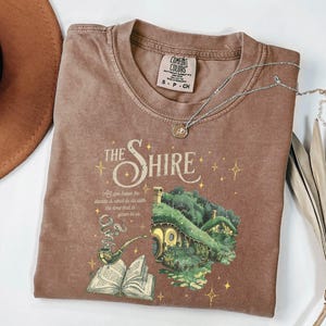 May include: A brown t-shirt with a vintage-style graphic of a hobbit house and a quote from The Lord of the Rings. The text reads "The Shire" and "All we have to decide is what to do with the time that is given to us."