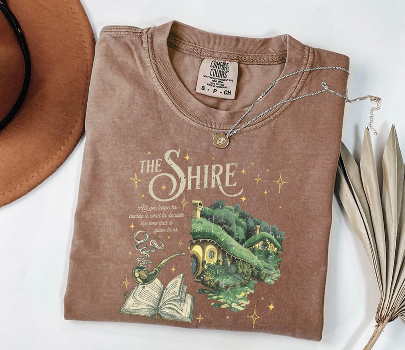 May include: A brown t-shirt with a vintage-style graphic of a hobbit house and a quote from The Lord of the Rings. The text reads "The Shire" and "All we have to decide is what to do with the time that is given to us."