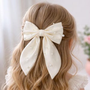 Può includere: Fiocco per capelli in raso avorio ornato di perle finte sparse. Il fiocco è legato in uno stile grande ed elegante ed è fissato tra i capelli. I capelli sono lunghi e mossi, di colore biondo chiaro. Un accessorio per capelli classico.