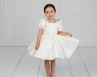 May include: A young girl wearing a white satin dress with a square neckline and puffed sleeves. The dress has a full skirt and is paired with matching ballet flats. The background is a white wall and floor.