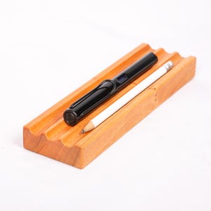 May include: A wooden pen and pencil holder with a wavy design. The holder is made of light brown wood and has three slots for pens or pencils. A black pen and a white pencil are in the holder.
