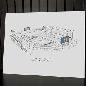 Puede incluir: Un boceto en blanco y negro del estadio de Michigan, tambi&eacute;n conocido como "The Big House". La obra de arte representa el exterior del estadio, incluyendo los asientos, los marcadores y el campo. El nombre y el apodo del estadio est&aacute;n escritos debajo.