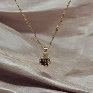 May include: A gold-coloured necklace with a pomegranate pendant. The pendant has a gold-coloured exterior and a red interior, suspended from a delicate chain. The necklace is displayed on a satin-like fabric.