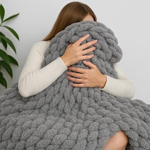 May include: A grey chunky knit throw blanket with a cable knit pattern. The blanket is soft and fluffy, and it is perfect for snuggling up on the sofa.