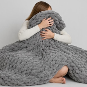 Può includere: Una coperta di maglia grossa di colore grigio con una persona seduta sopra. La coperta è realizzata in filato spesso e morbido e presenta un motivo a treccia.