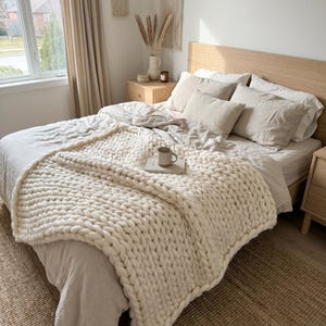 Puede incluir: Una escena de dormitorio acogedora con una cama con una manta de punto grueso de color crema. La cama está hecha con ropa de cama y almohadas de tonos neutros. Una taza y un libro descansan sobre la manta.