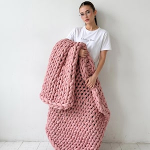 Puede incluir: Una manta grande de punto grueso en color rosa suave. La manta se sostiene, mostrando su textura gruesa y tejida. La manta parece estar hecha de un material suave y afelpado, perfecto para acurrucarse. Las dimensiones de la manta no son visibles.