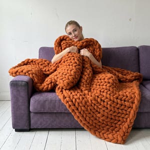 Könnte beinhalten: Eine große, grob gestrickte Decke in einem warmen Orangeton. Die Decke ist über ein lila Sofa drapiert und schafft eine gemütliche und einladende Szene. Das dicke Garn ist sichtbar und zeigt die Textur und das Gewicht der Decke. Die Decke misst etwa 152 cm x 203 cm.