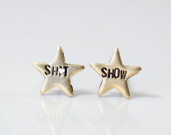 May include: A pair of gold star-shaped earrings with the words "SH*T" and "SHOW" engraved on them.