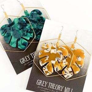May include: Two pairs of gold hoop earrings with a large, green and gold, or yellow and white, marbled monstera leaf design. The earrings are displayed on black cards with the text "GREY THEORY MILL" in gold lettering.