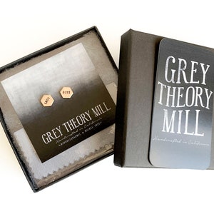 May include: A black gift box with a white card that says "GREY THEORY MILL Handcrafted in California". Inside the box is a white card with the words "GREY THEORY MILL Handcrafted in California Hypoallergenic & Nickel Free" and two gold hexagonal earrings with the words "BOSS" and "LADY" engraved on them.