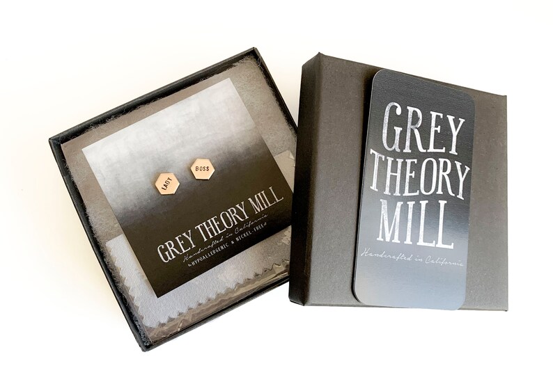 May include: A black gift box with a white card that says "GREY THEORY MILL Handcrafted in California". Inside the box is a white card with the words "GREY THEORY MILL Handcrafted in California Hypoallergenic & Nickel Free" and two gold hexagonal earrings with the words "BOSS" and "LADY" engraved on them.