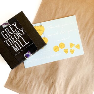 May include: A brown paper bag with a small black box and a thank you card. The box has a sticker that says "GREY THEORY MILL Handmade in California". The card has a blue background with white text that says "Thank you so much for your order!" and a website address: "greytheorymill@gmail.com".