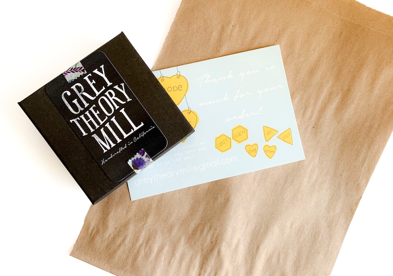 May include: A brown paper bag with a small black box and a thank you card. The box has a sticker that says "GREY THEORY MILL Handmade in California". The card has a blue background with white text that says "Thank you so much for your order!" and a website address: "greytheorymill@gmail.com".