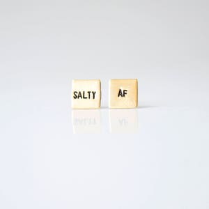 May include: Two small gold-colored square earrings with the words "SALTY" and "AF" engraved on them.