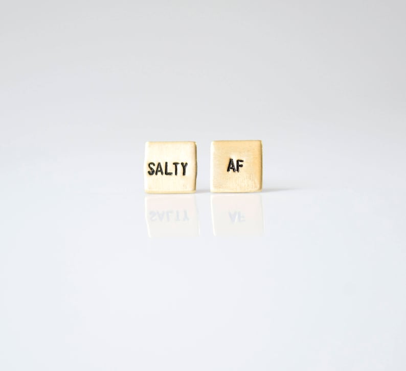 May include: Two small gold-colored square earrings with the words "SALTY" and "AF" engraved on them.