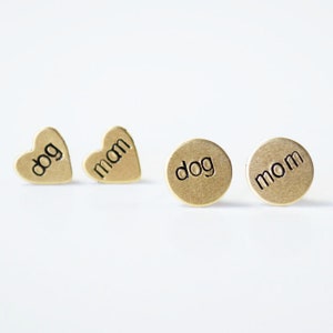 May include: Four gold-toned earrings with the words "dog" and "mom" engraved on them. Two earrings are heart-shaped and two are round.
