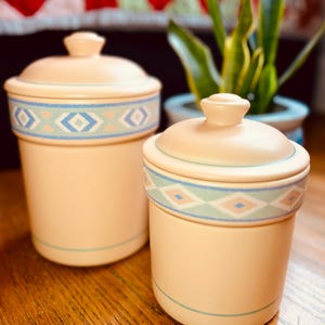 May include: Two ceramic canisters with lids, both featuring a light pink base with a blue and green geometric pattern around the middle. The canisters are on a wooden surface.